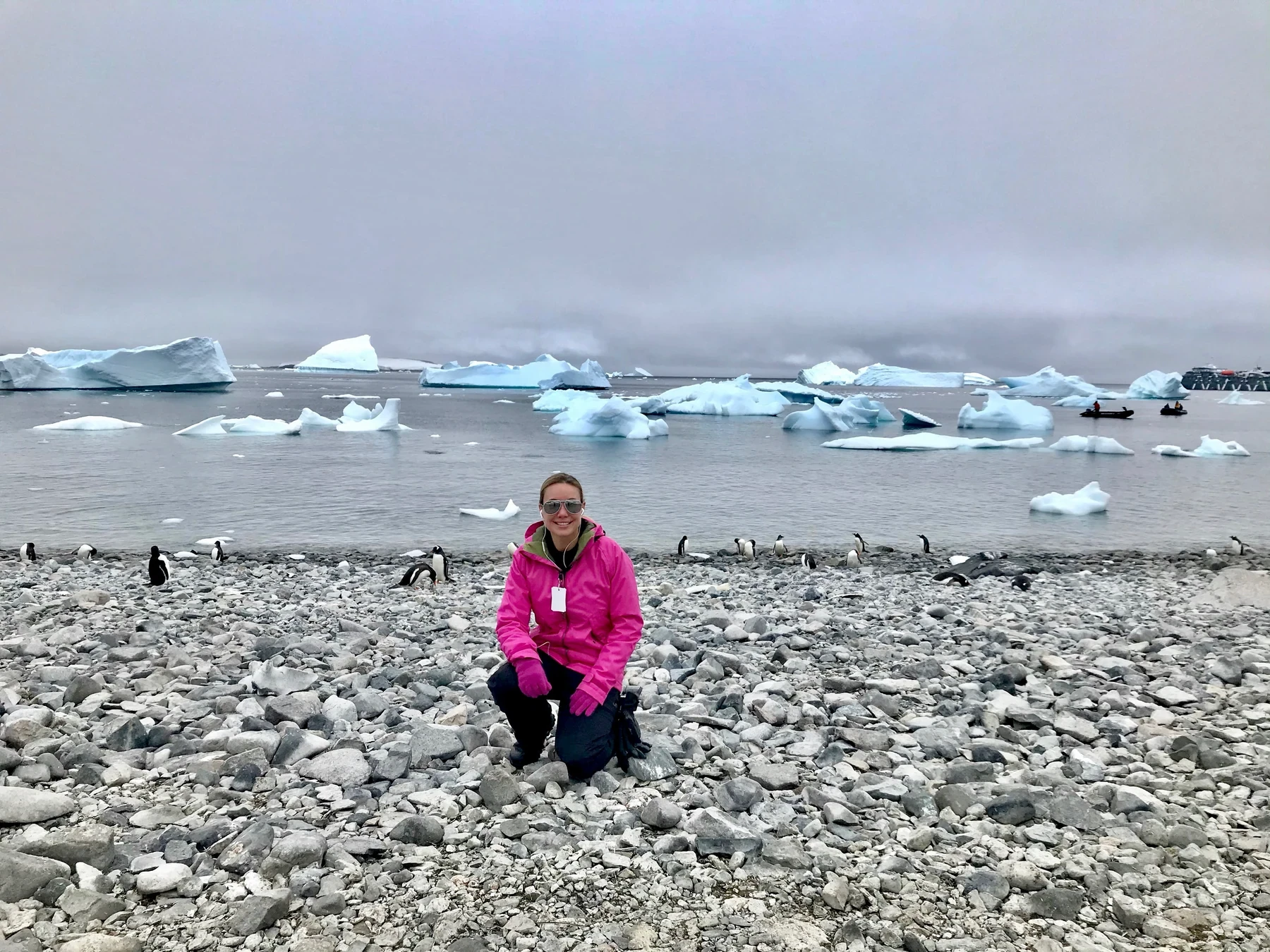 Cassandra Mann kneeling on an Antarctic beach with penguins and icebergs behind her.